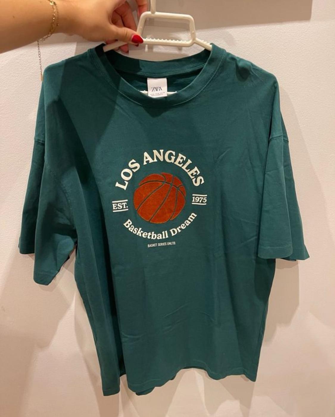 Green T-shirt with basketball design and text "LOS ANGELES Basketball Dream EST. 1975". Hand holding the hanger.