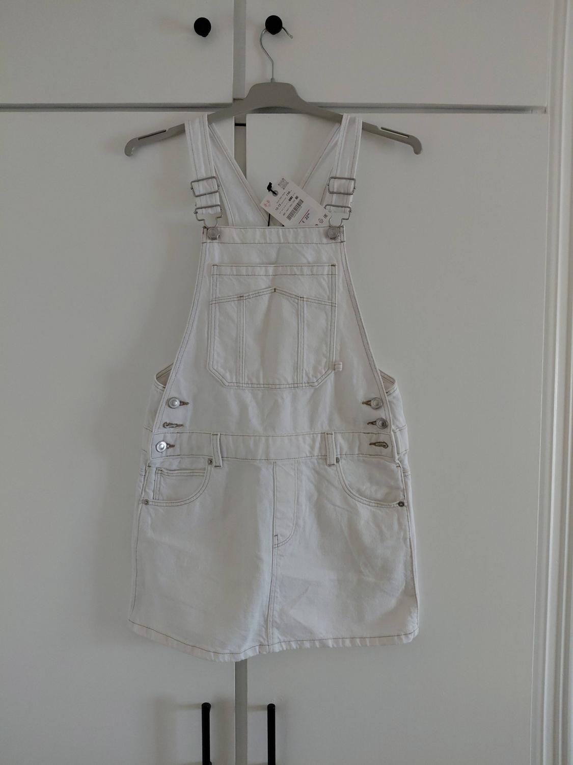 White denim dungaree skirt with metal buttons and a front pocket, hanging on a hanger over the doors of a white wardrobe.