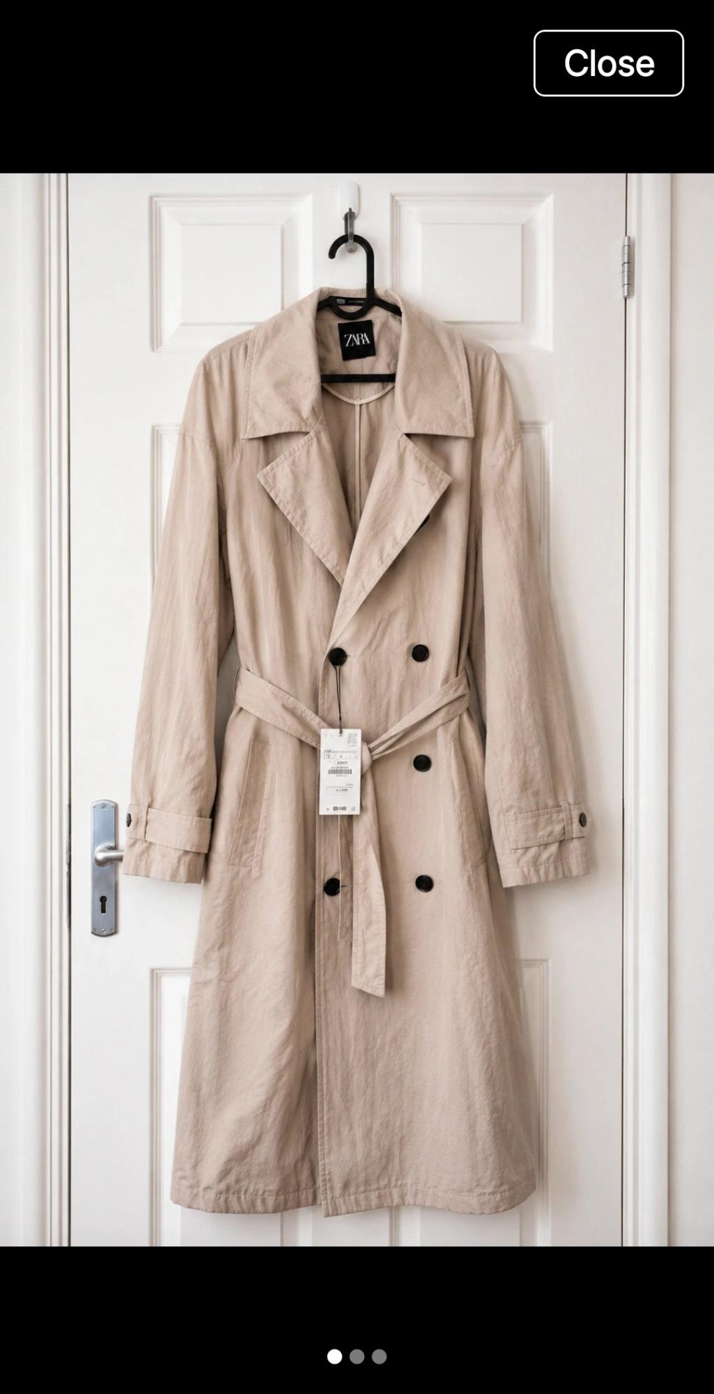 Double-breasted beige trench coat with dark buttons and belt, hanging on a white door. ZARA label. Text: Close.