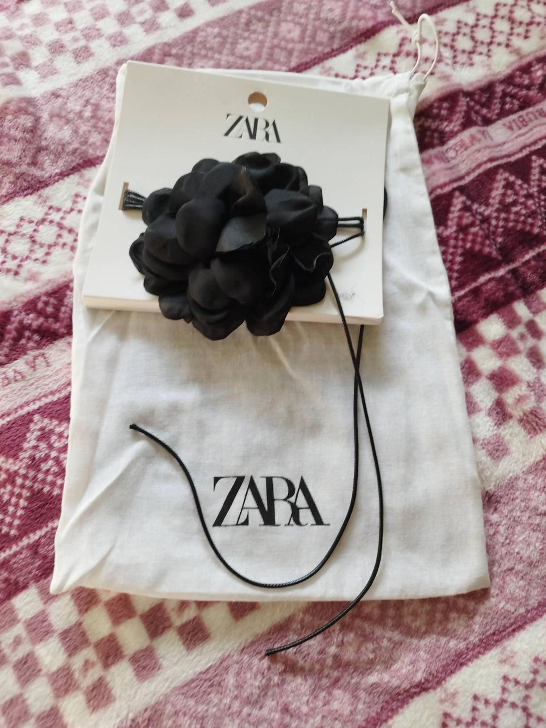 Black fabric flower with a cord on a white tag and bag, both featuring the ZARA text. Relaxed composition on a pink patterned blanket.