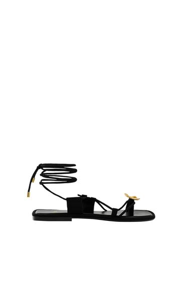 Black strappy sandals with a gold detail on the front and long laces to tie at the ankle, on a white background.