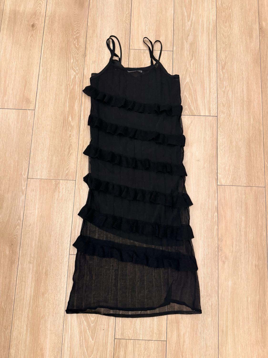 Semi-sheer black dress with thin straps and horizontal ruffles, on a light wooden floor.
