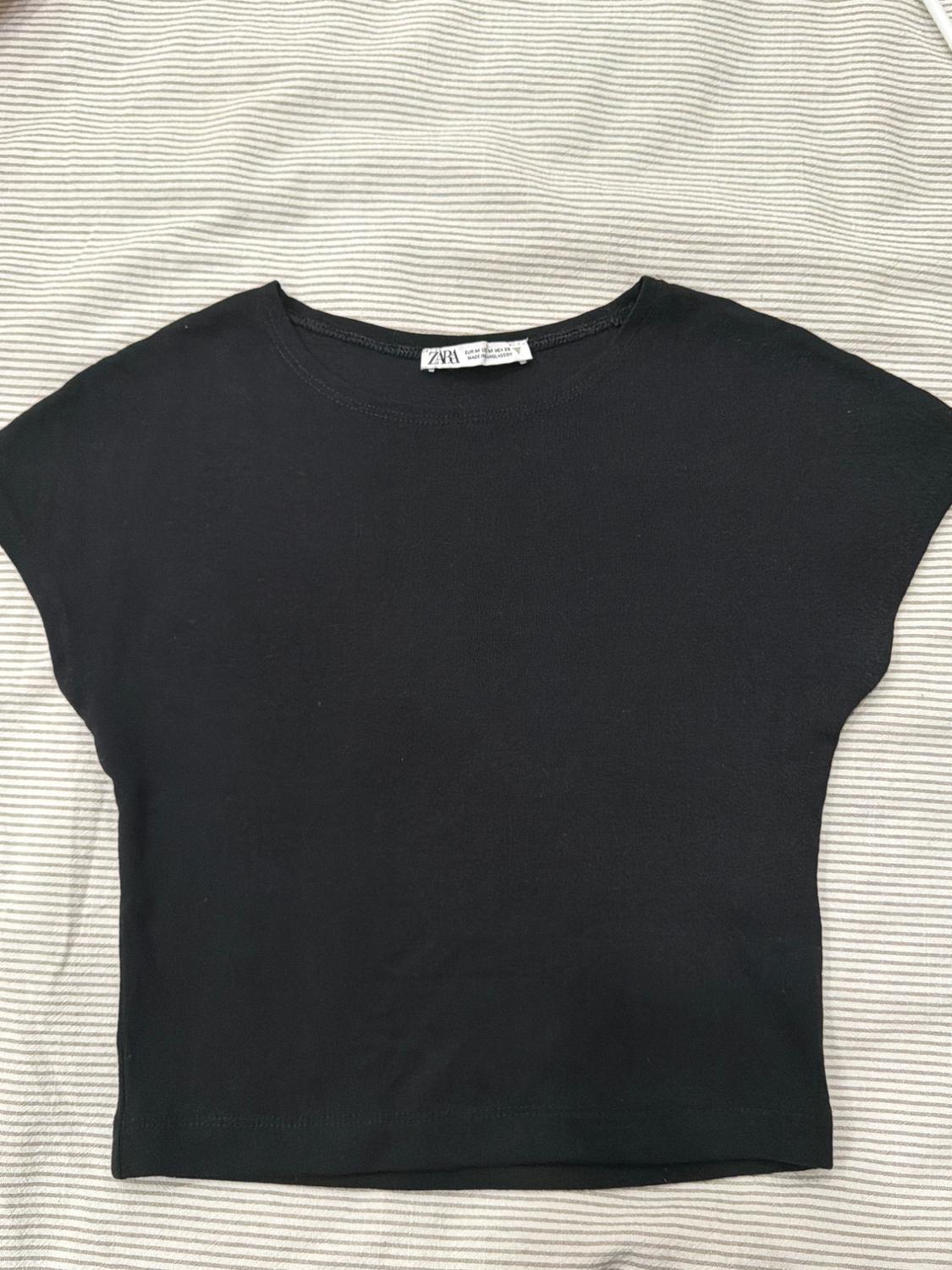 Black short-sleeved t-shirt on a striped fabric background. Visible label on the collar with text "ZARA".