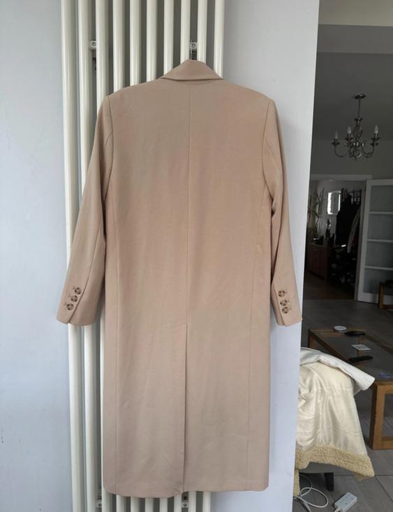 Beige coat hanging on a white radiator, with buttons on the sleeves. Living room background with chandelier and furniture.