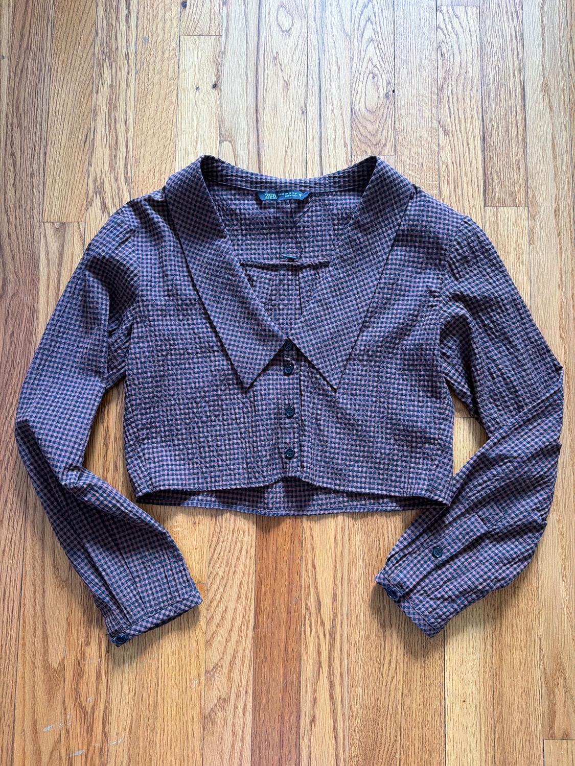 Brown and black checked cropped shirt with a wide collar and front buttons, on a light wooden floor.