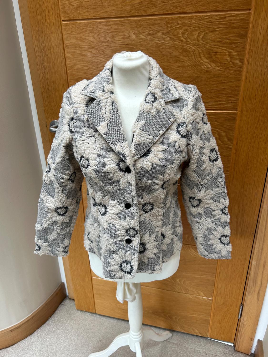 Grey textured jacket with black buttons and a pattern of large raised white flowers on a mannequin in front of a wooden door.