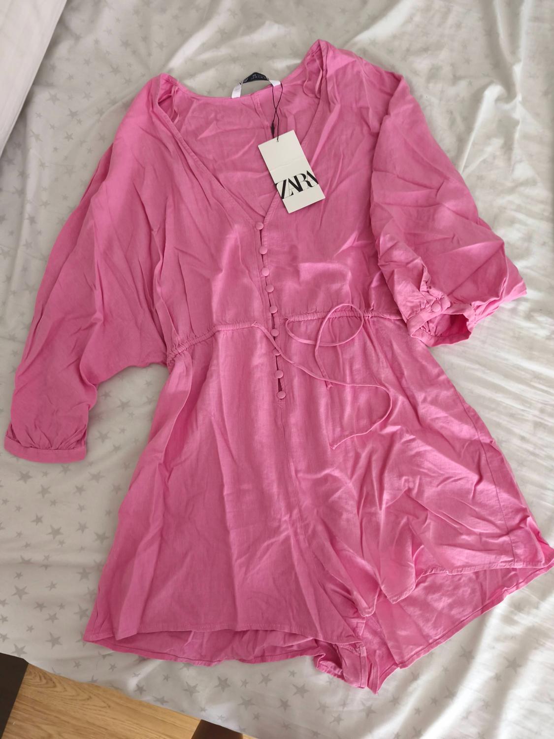 Pink romper with puff sleeves and front buttons, on a bed with star-print sheets. Visible label with text "ZARA".