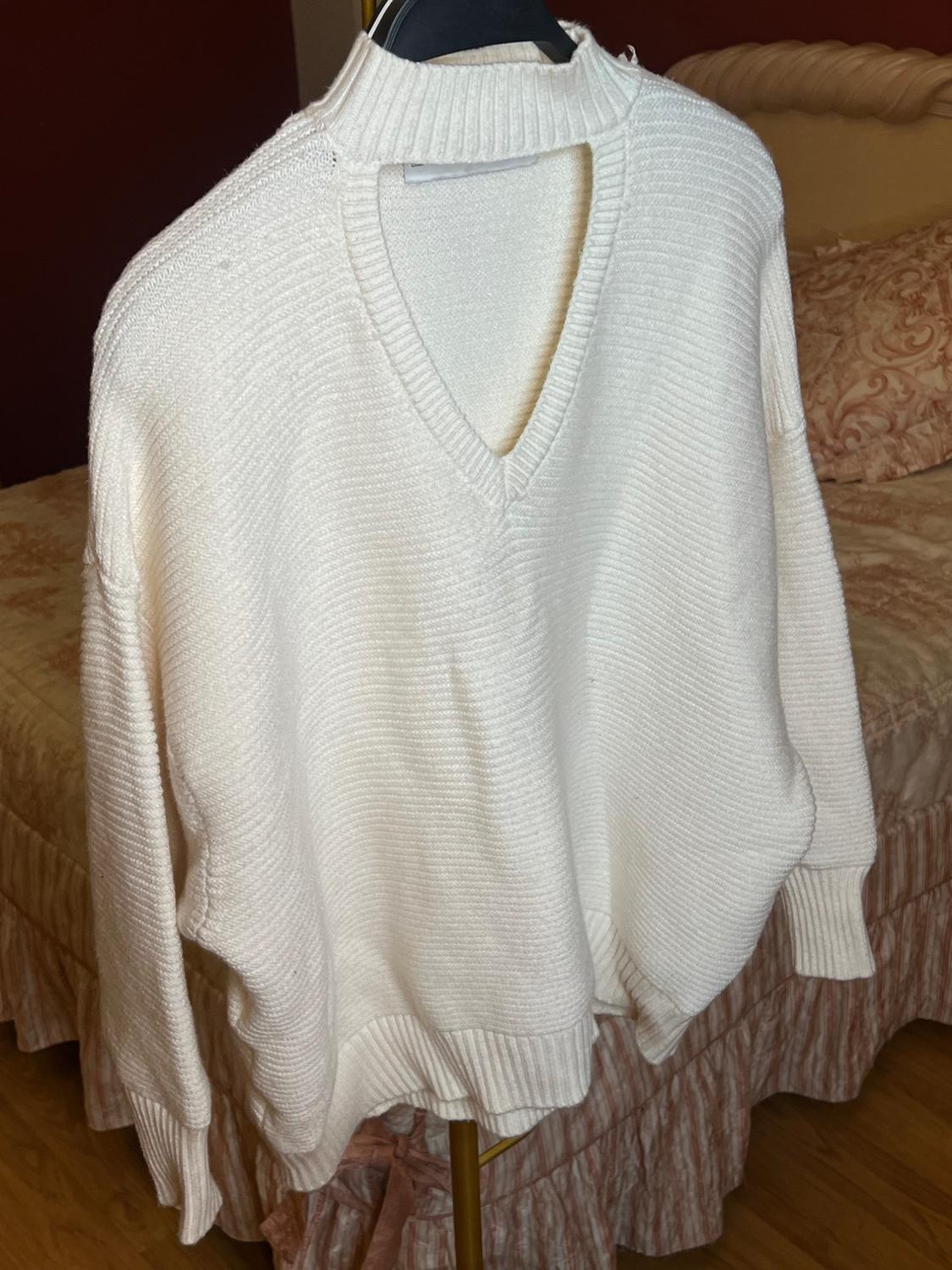 White chunky knitted jumper with a high neck and an opening at the back, hanging on a hanger over a bed with a beige bedspread.