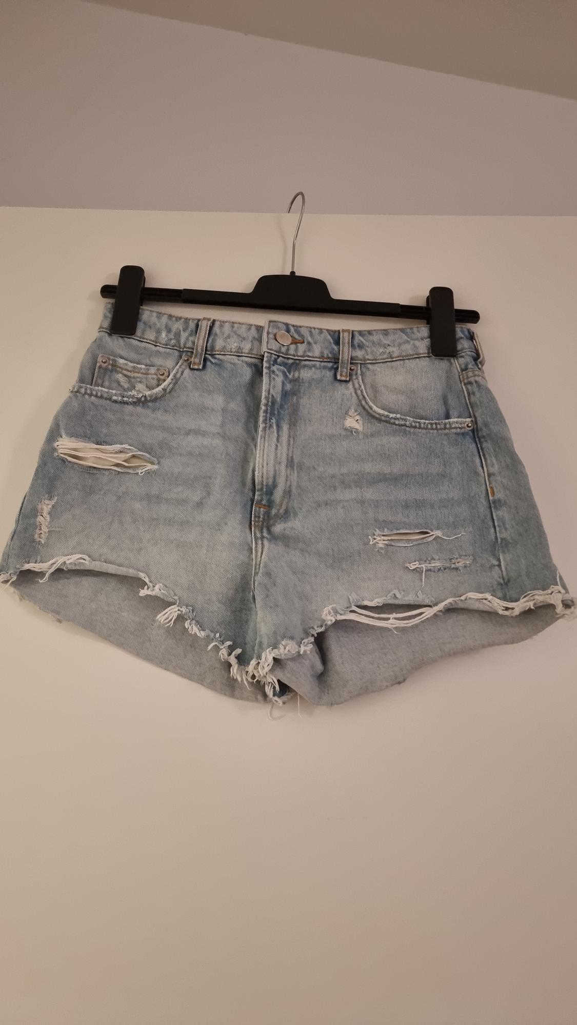 Distressed denim shorts hanging on a black hanger against a white wall. They feature rips and frayed edges.