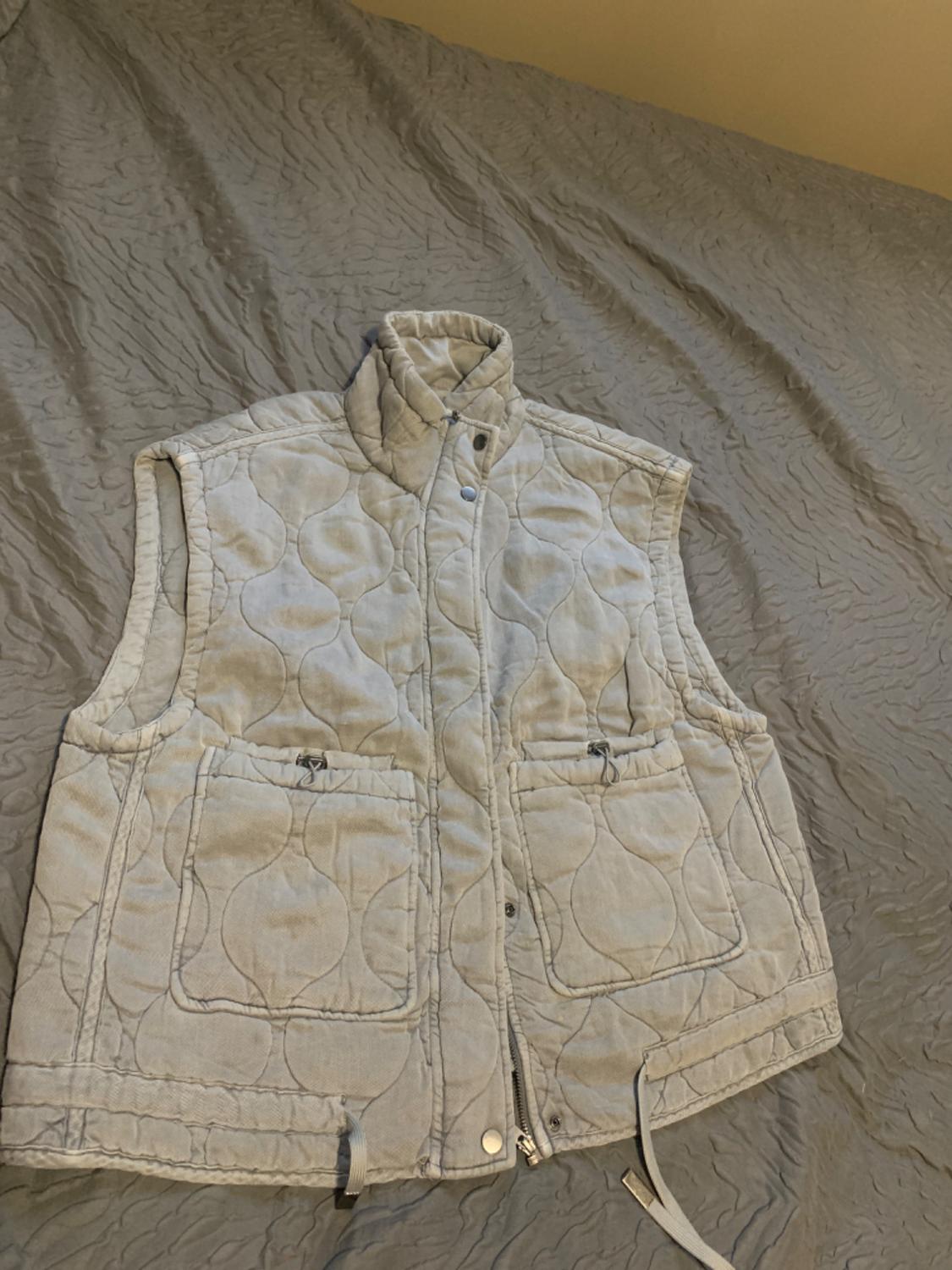 Light grey padded gilet with large pockets and zip fastening, over a grey bedspread with a wavy texture.