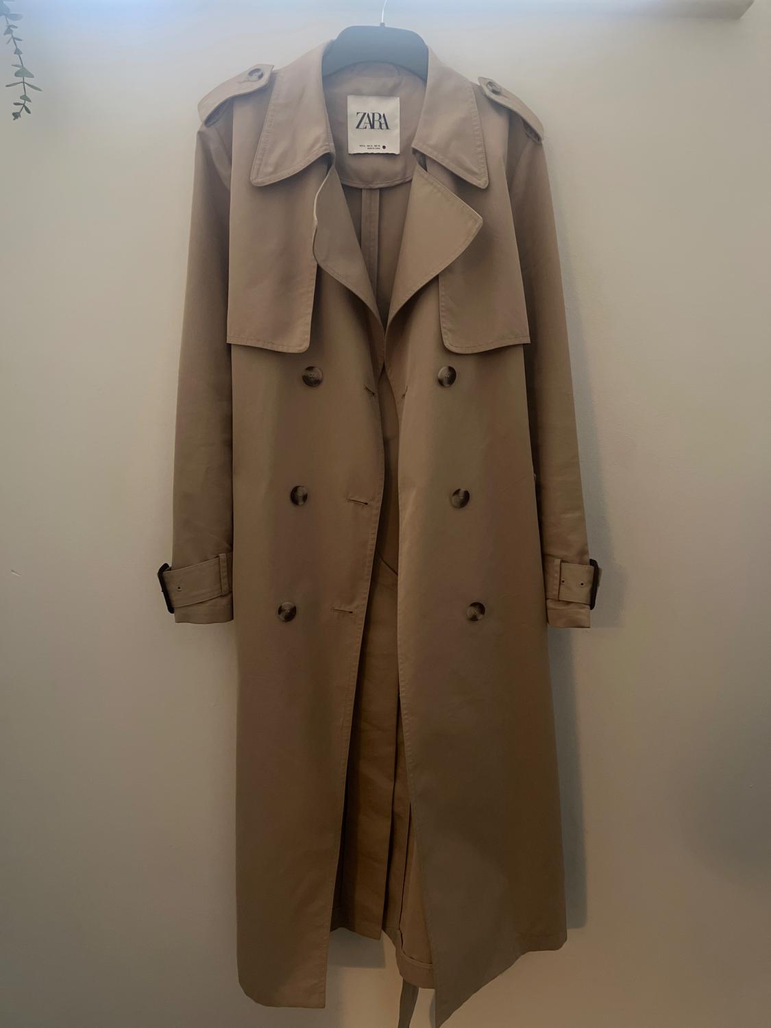 Beige trench coat hanging on a hanger with a visible label that says "ZARA". White background and soft light.