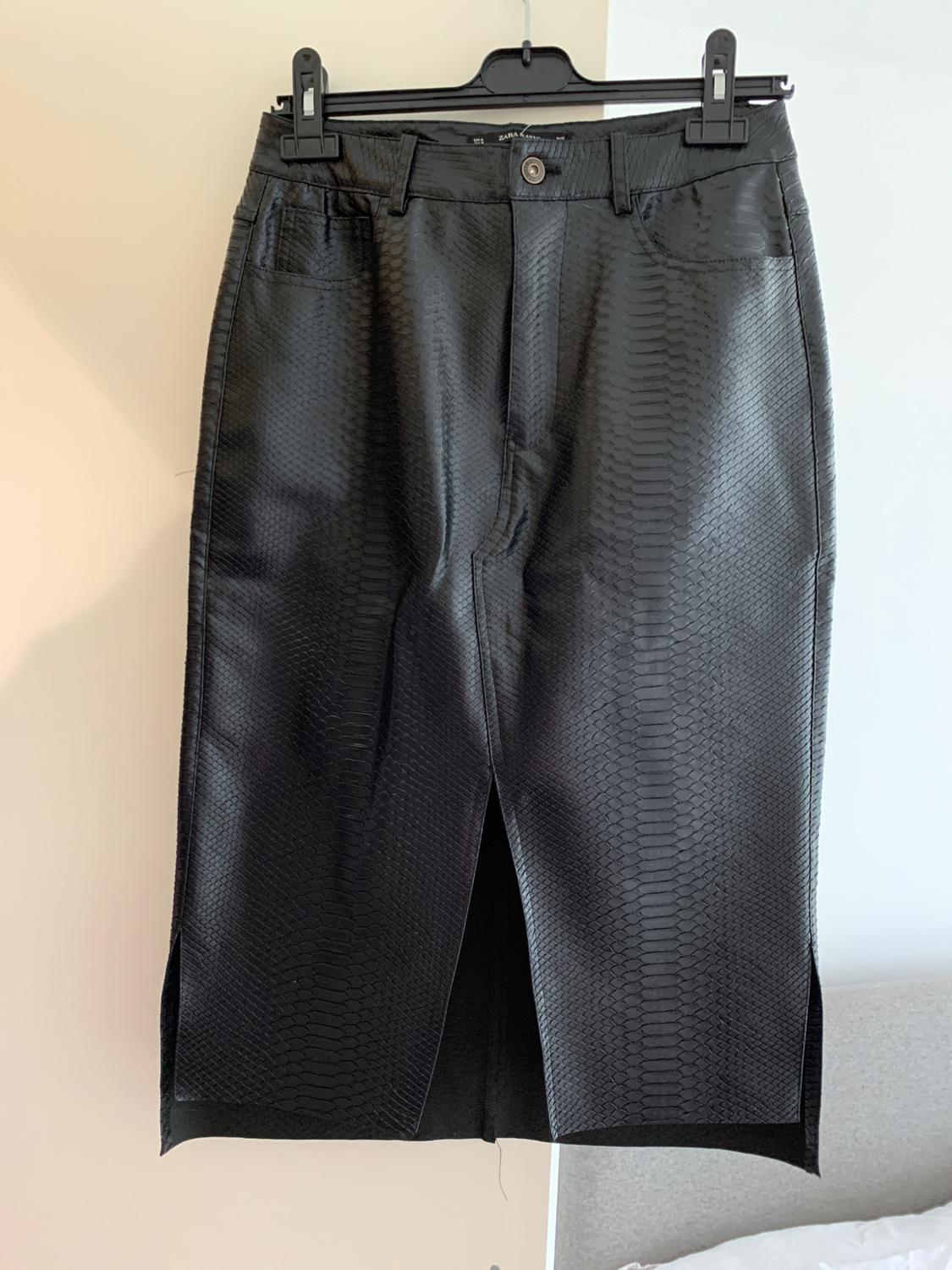 Black reptile-textured skirt hanging on a black hanger, with front slit and pockets, on a light background.