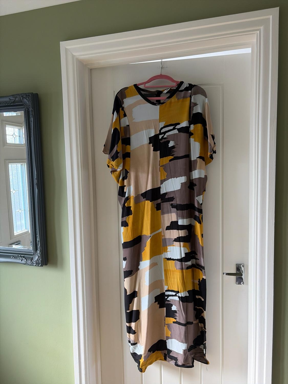 Mustard, black and beige abstract print maxi dress, hanging on a pink hanger on a white door.