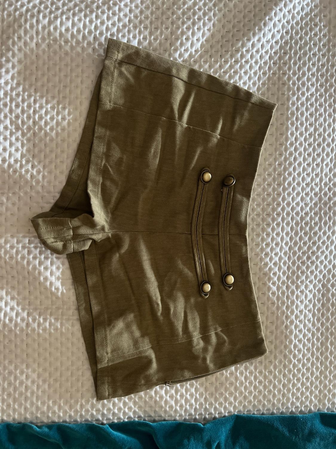 Olive green shorts with straps and decorative metallic buttons on the front, on a textured white surface.