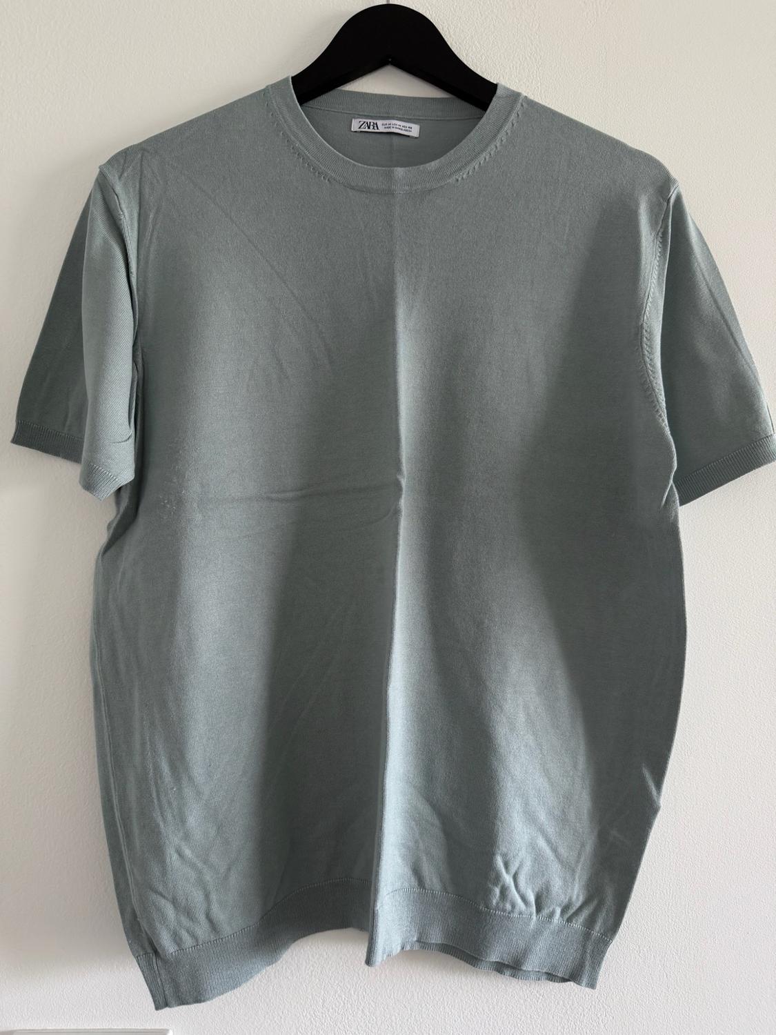 Blue-gray short-sleeve knit T-shirt hanging on a black hanger against a white background. The label reads ZARA.