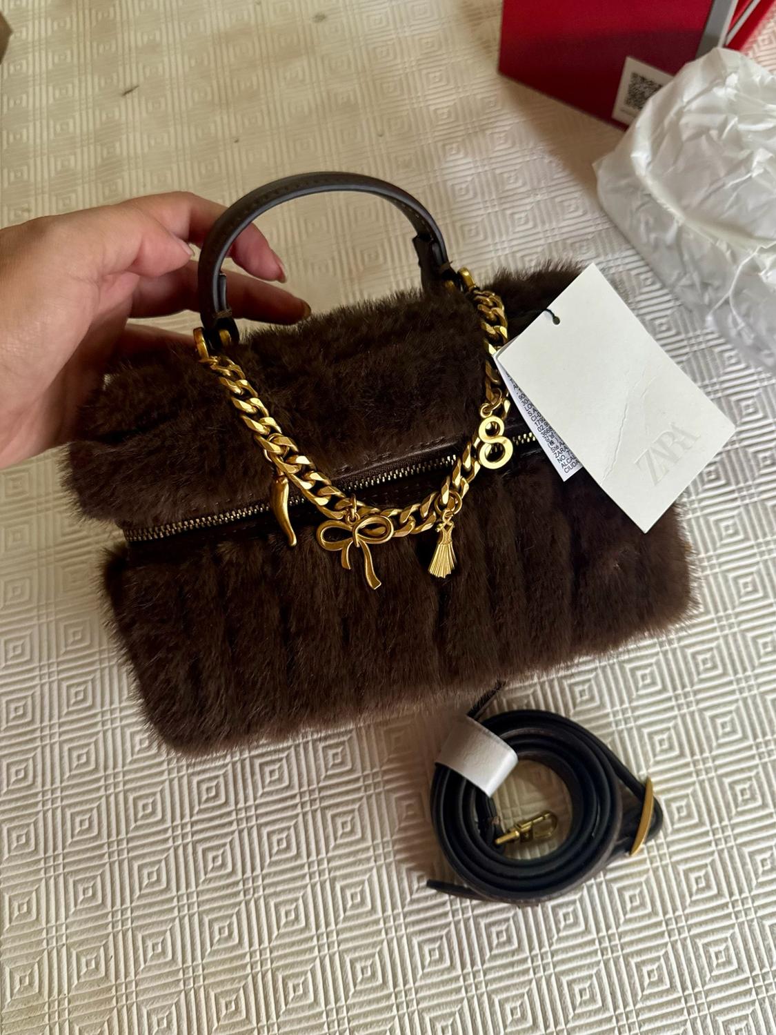 Hand holding a brown furry handbag with a handle, gold chain, charms and a ZARA label. Coiled strap beside it on a white surface.