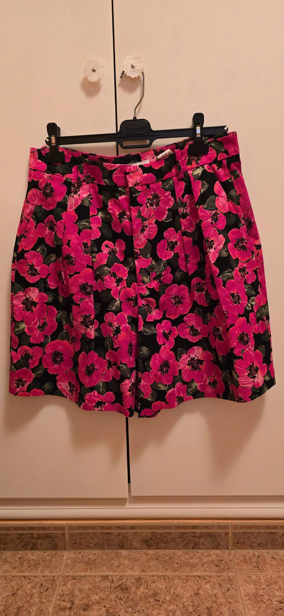 Pleated shorts with a large pink floral print on a black background, hanging on a hanger in front of a white wardrobe.