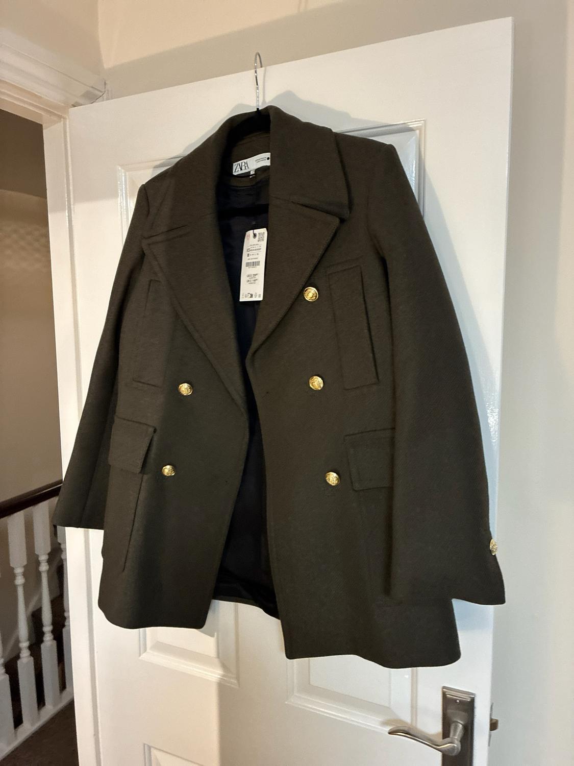 Green double-breasted coat with gold buttons and a ZARA label, hanging on a white door in an interior with a staircase in the background.