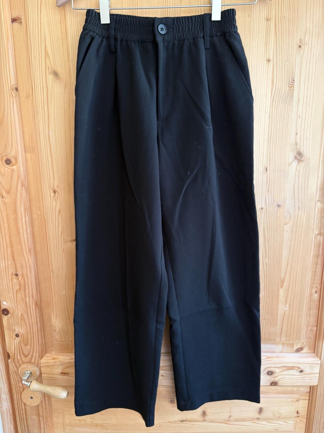 Black wide-leg trousers with an elasticated waistband and button, hanging on a hanger against a light wood door. Relaxed style.