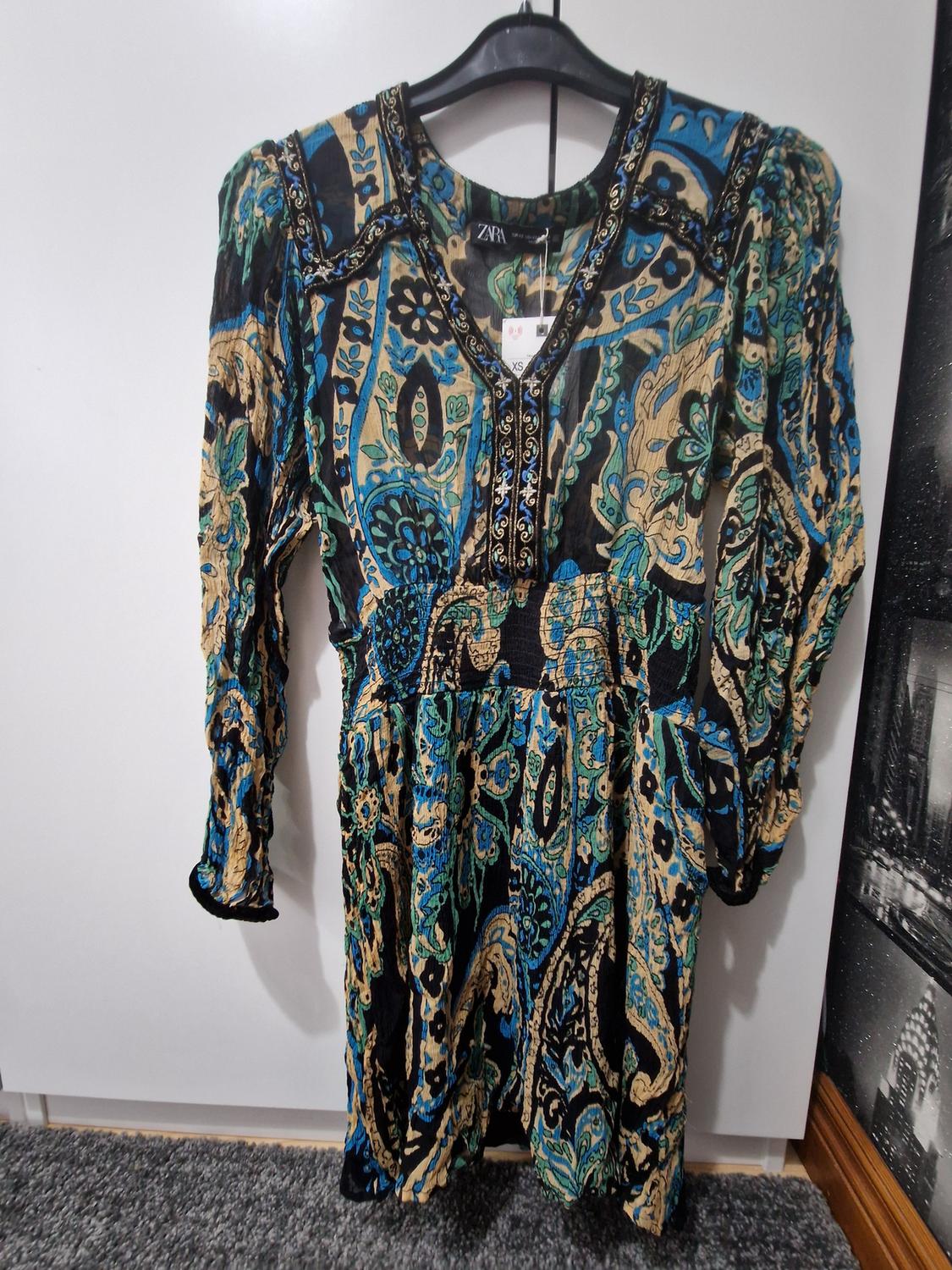 Long-sleeved dress with a blue, black and beige print, V-neck and elasticated waist, hanging on a hanger. Text ZARA XS.