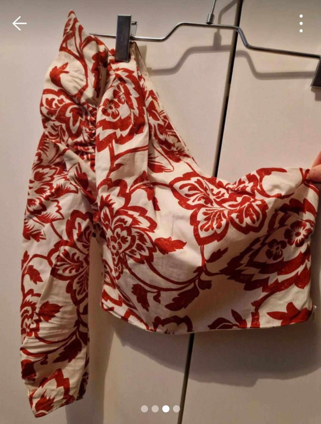 Red floral print one-shoulder blouse on a white background, hanging on a metal hanger in front of a wardrobe.