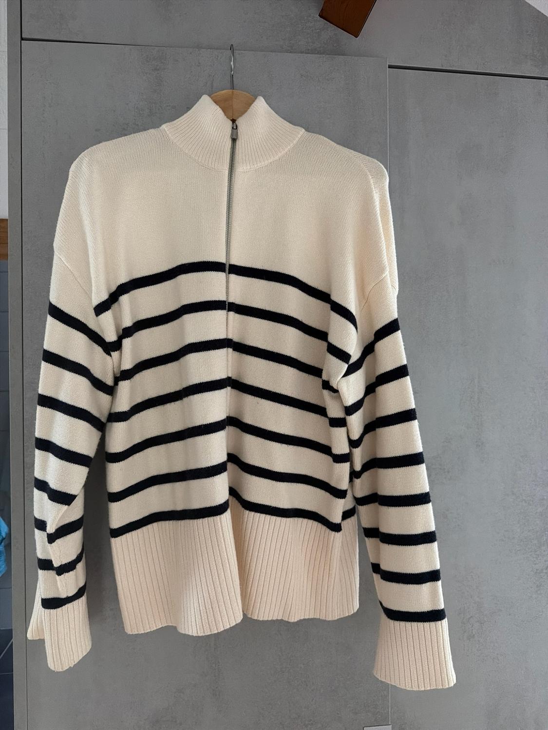 Cream knitted jumper with black stripes and a high neck with a zip, hanging on a wooden hanger against a grey background.