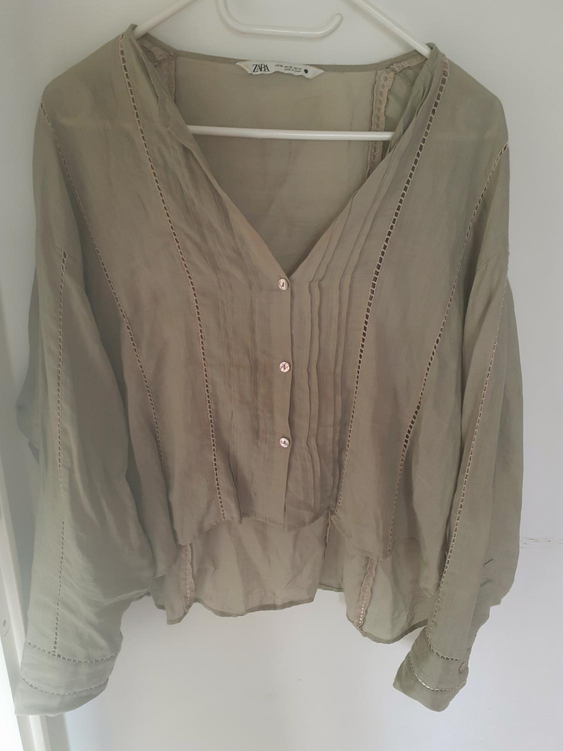 Olive green blouse on a white hanger. Long sleeves, V-neck, front buttons, pleats and openwork stitch details. Text: ZARA.