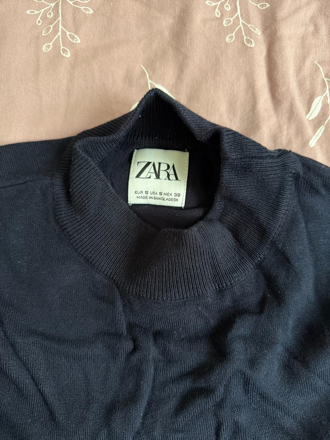 Dark blue knit turtleneck sweater on a printed pink background. Text: ZARA EUR S USA S MEX 38 MADE IN BANGLADESH.
