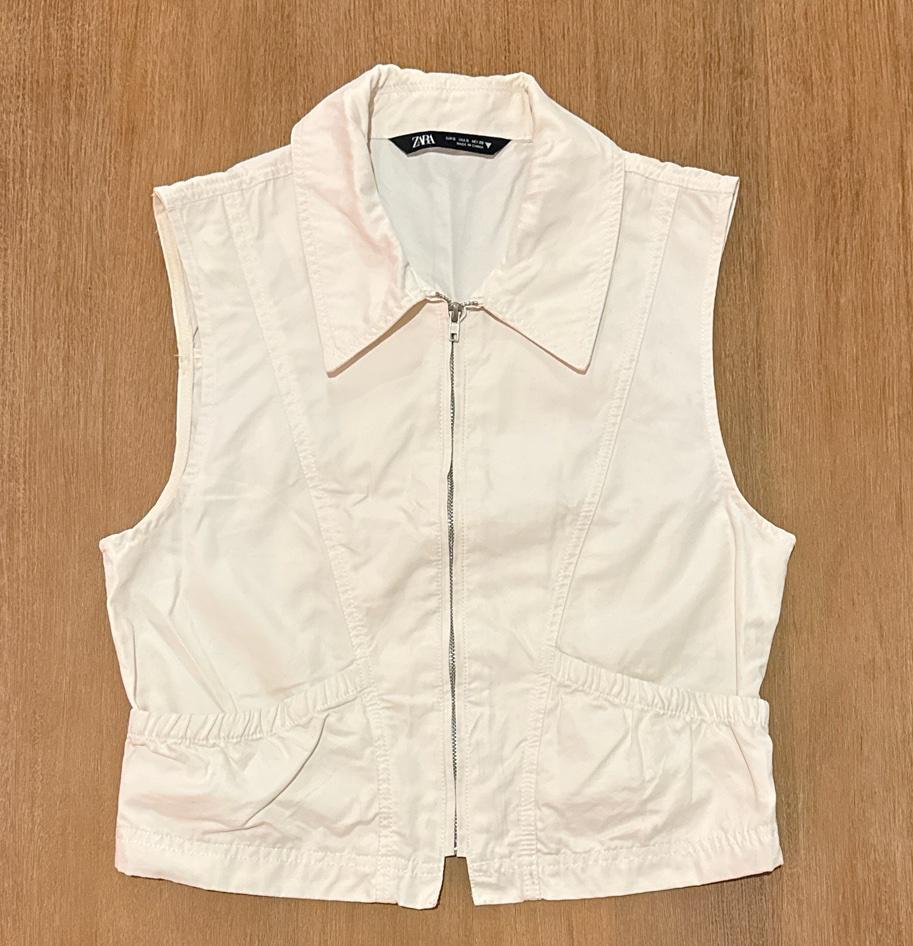 White vest with front zipper, collar, and gathered detail at the waist, on a wooden surface. ZARA interior label.