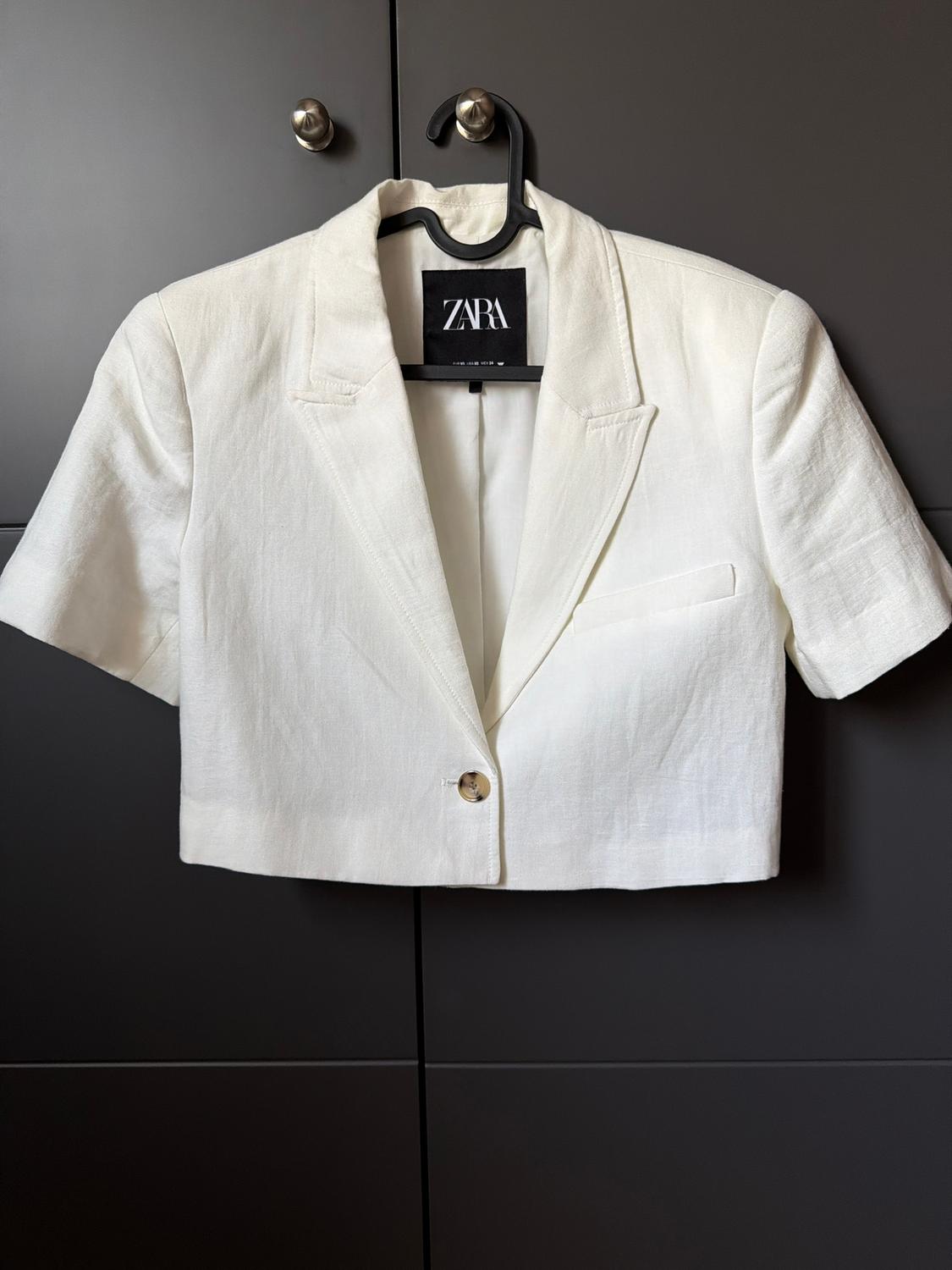 Short white, short-sleeved jacket hanging on a black hanger, with a visible Zara label, against a dark background.