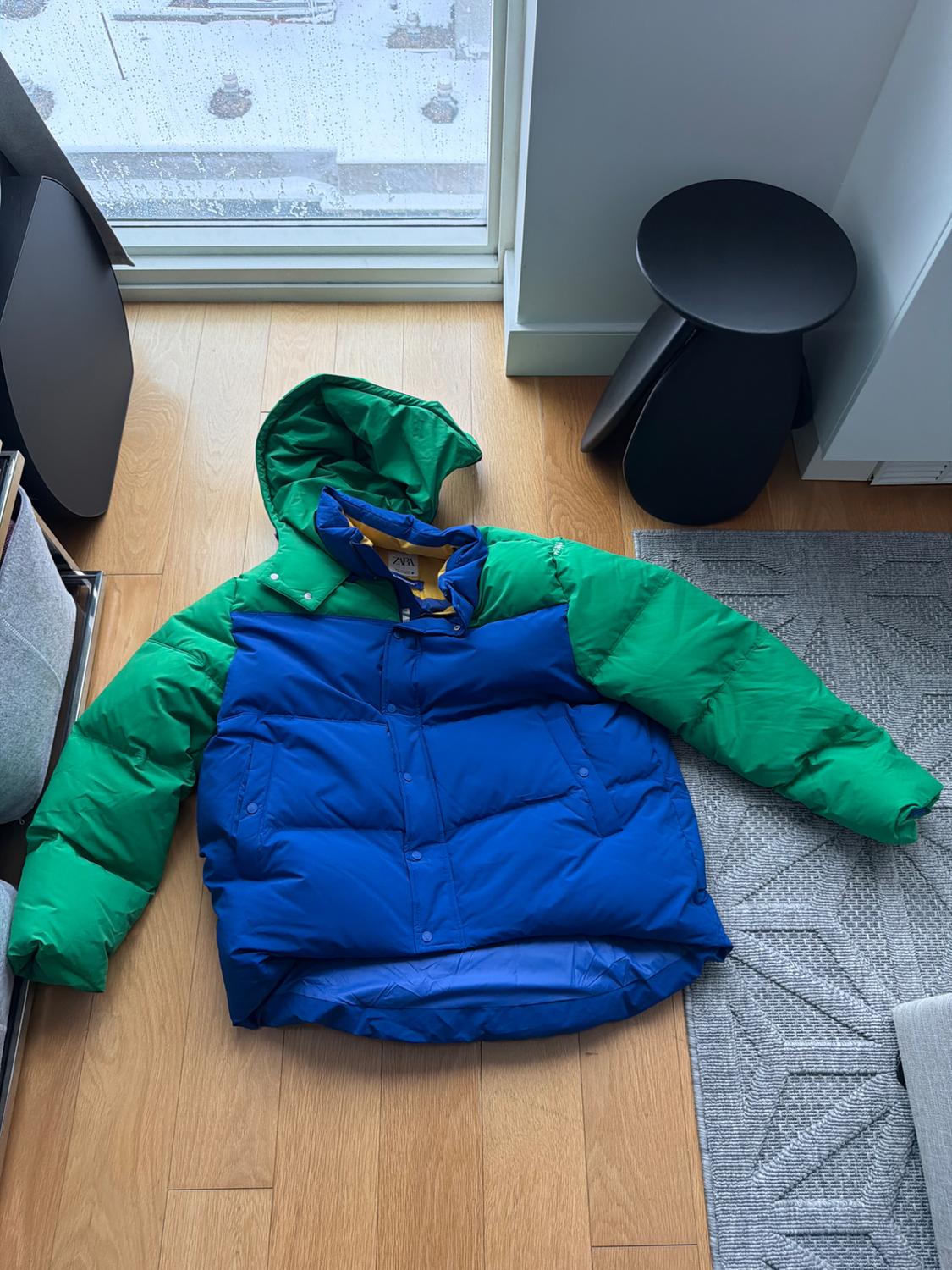 Blue and green padded jacket with a hood on a wooden floor next to a window with rain. Background with a gray carpet and a black stool.