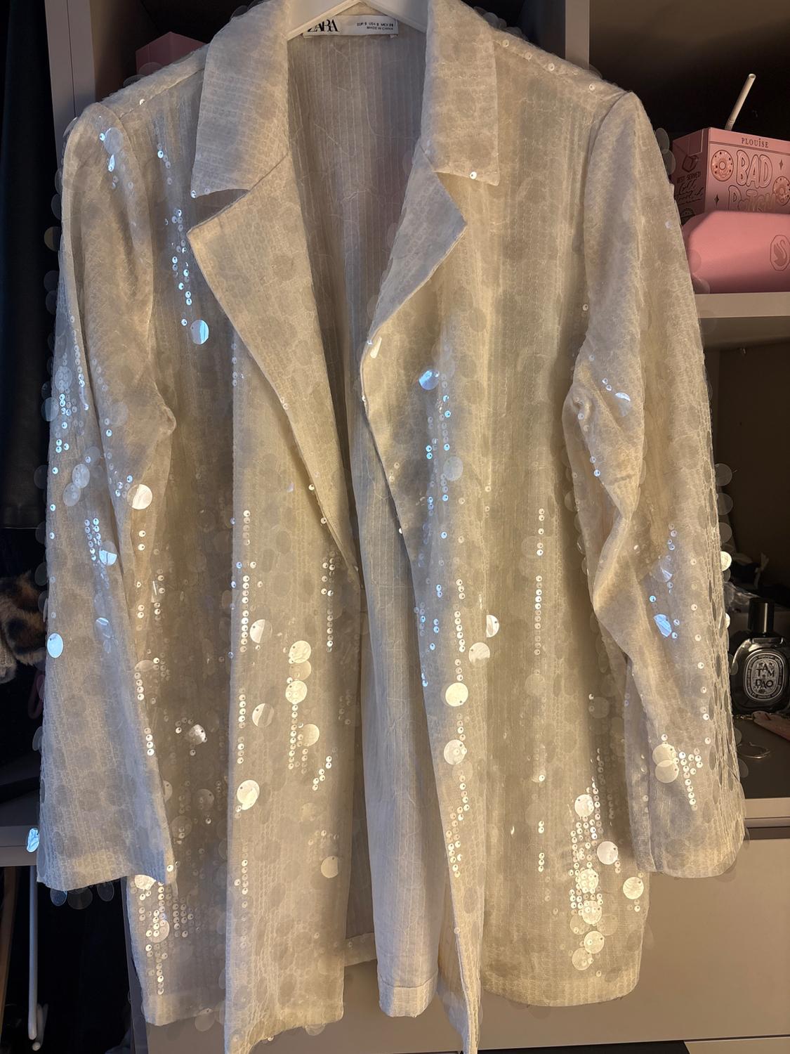 Jacket hanging in a wardrobe, light-coloured with shiny sequins. Label visible on the collar. Background with boxes and objects.