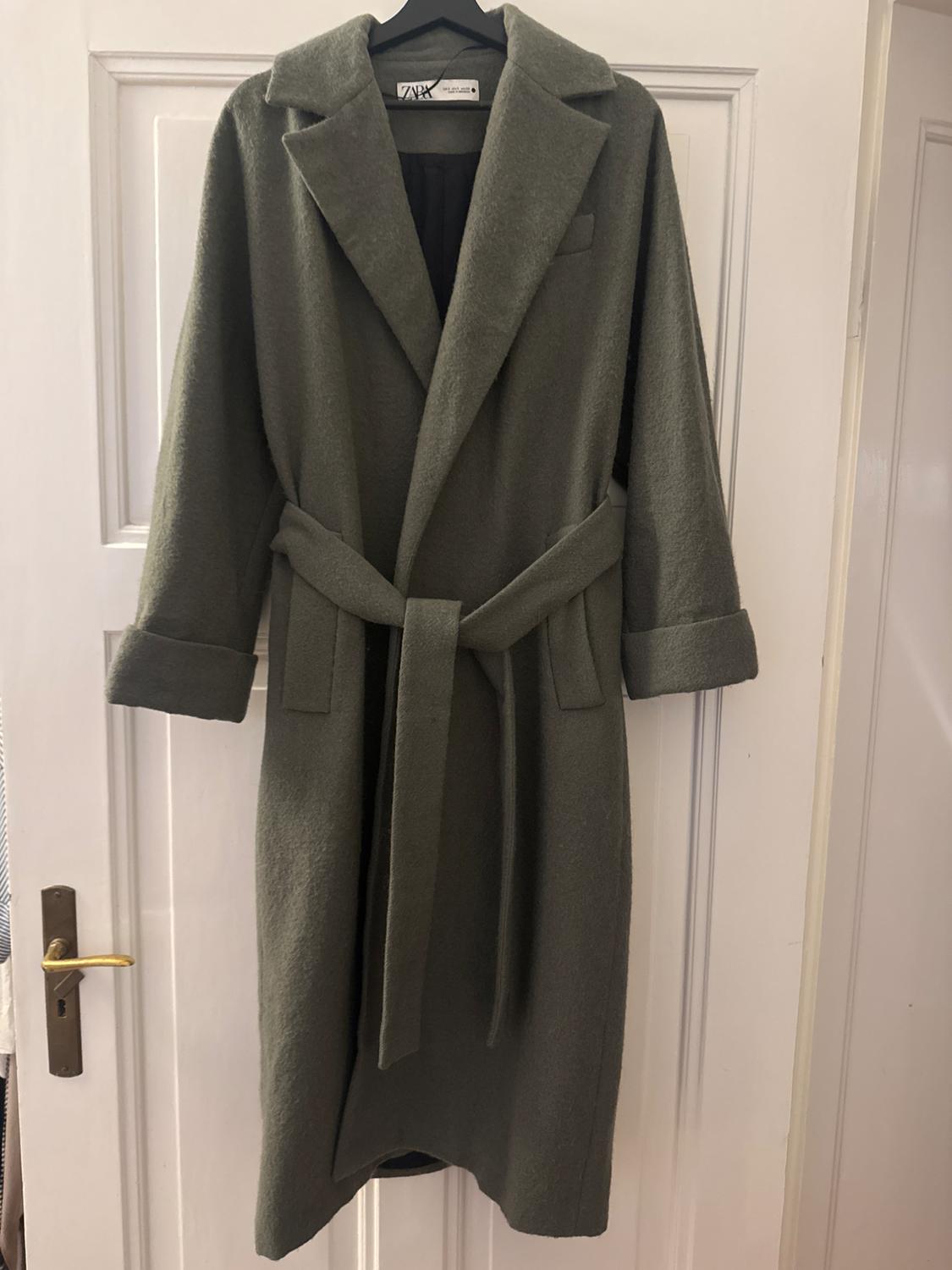 Olive green coat with belt hanging on a white door. Label visible on the collar with text "Zara".