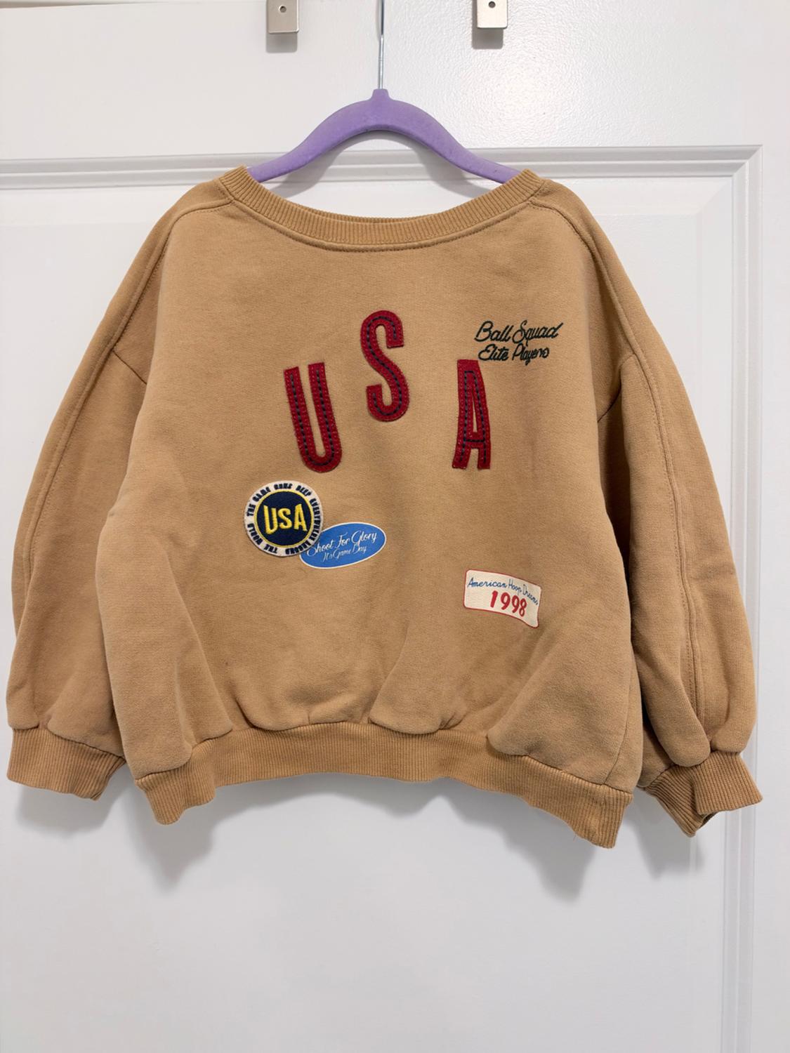 Beige sweatshirt hanging up with red USA lettering and patches with text: Ball Squad Elite Players, Shoot For Glory and American Hoop Dreams 1998.
