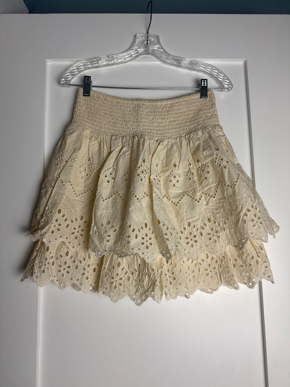 Cream short skirt with an elasticated waist and broderie anglaise ruffle design, hanging on a hanger over a white door.