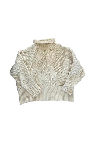 Chunky cream knit sweater with a high neck and cable-knit design. Long sleeves and ribbed cuffs. White background.