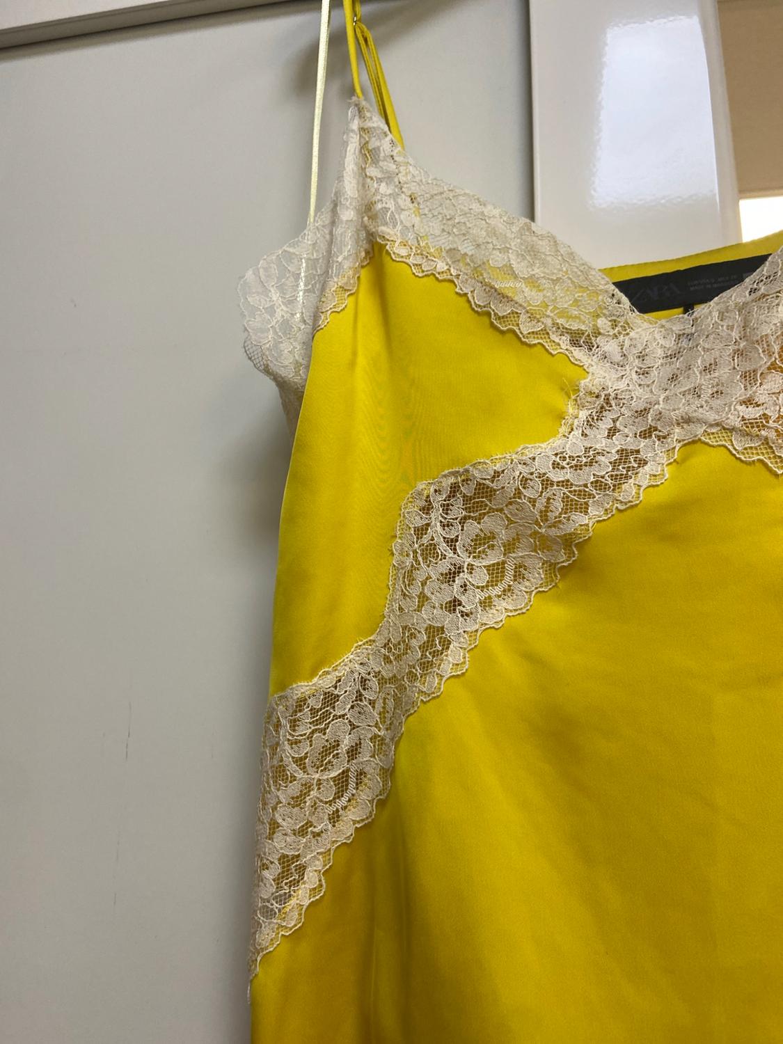 Yellow lingerie garment with thin straps and white lace at the neckline and crossing the chest, hanging on a white door. ZARA label.
