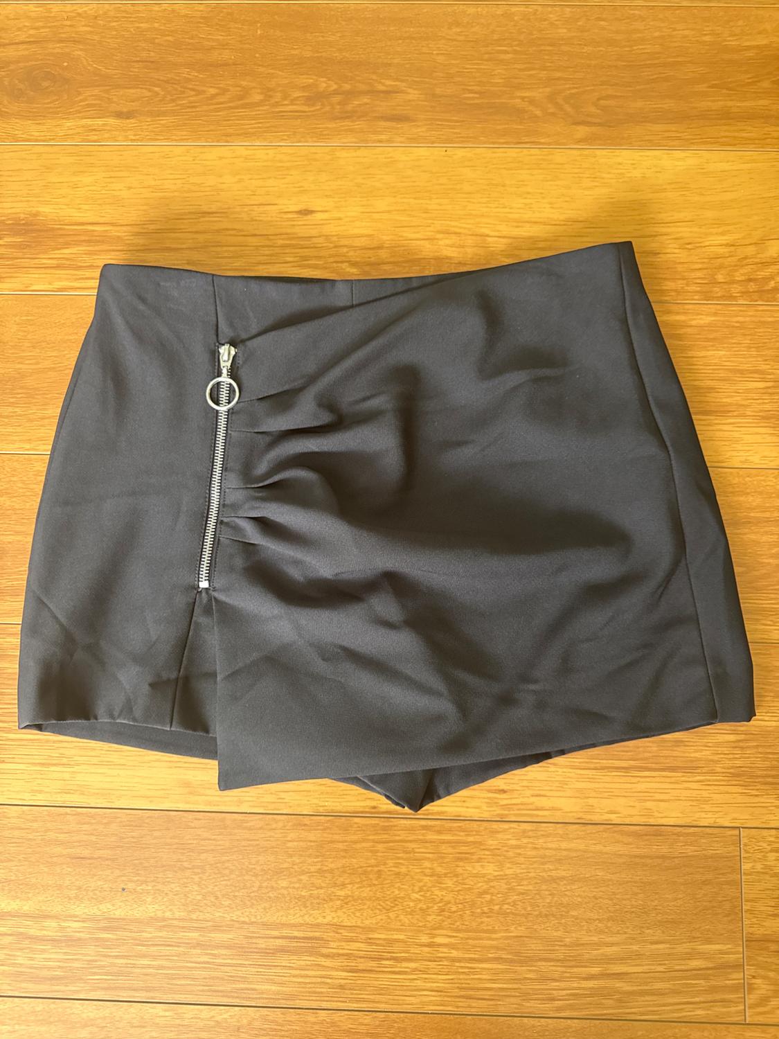 Black mini skirt with a side zip and ruched detail, on a light wooden floor.