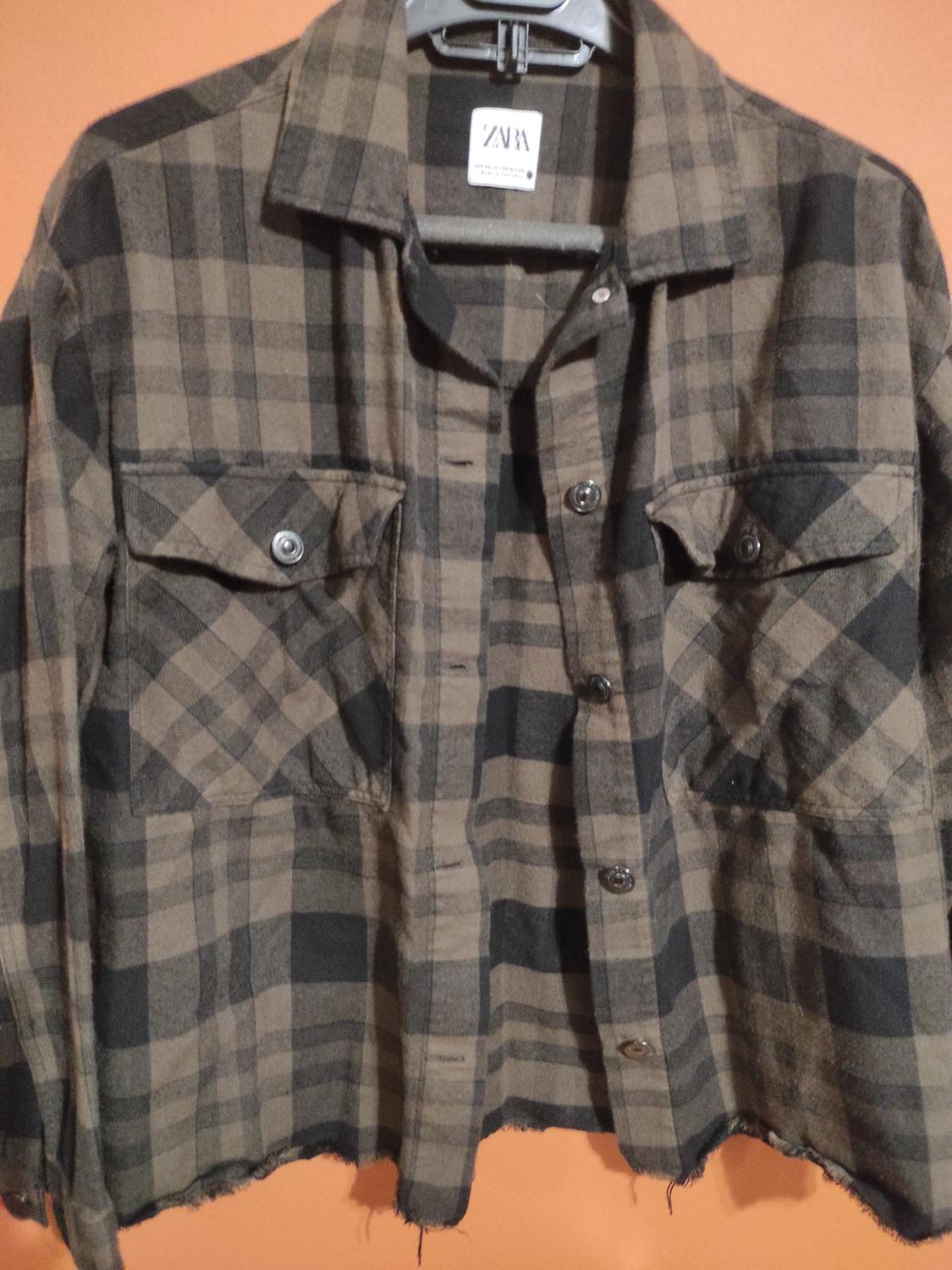 Brown and black checked shirt with front pockets and buttons, hanging on a hanger against an orange background. Visible tag.