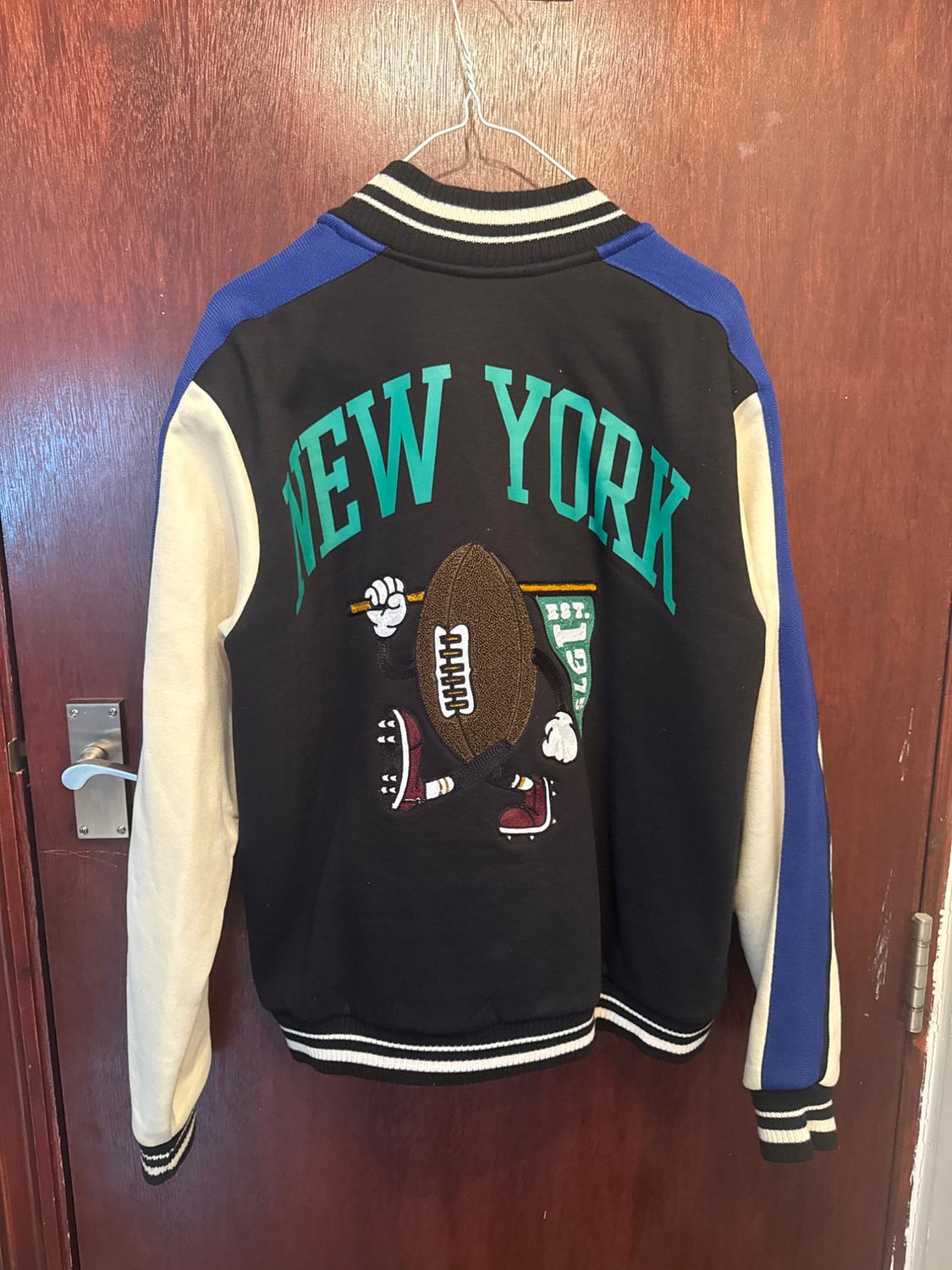 Black jacket with white and blue sleeves hanging on a door. Its back displays the text NEW YORK, EST. 1975 and an animated ball.