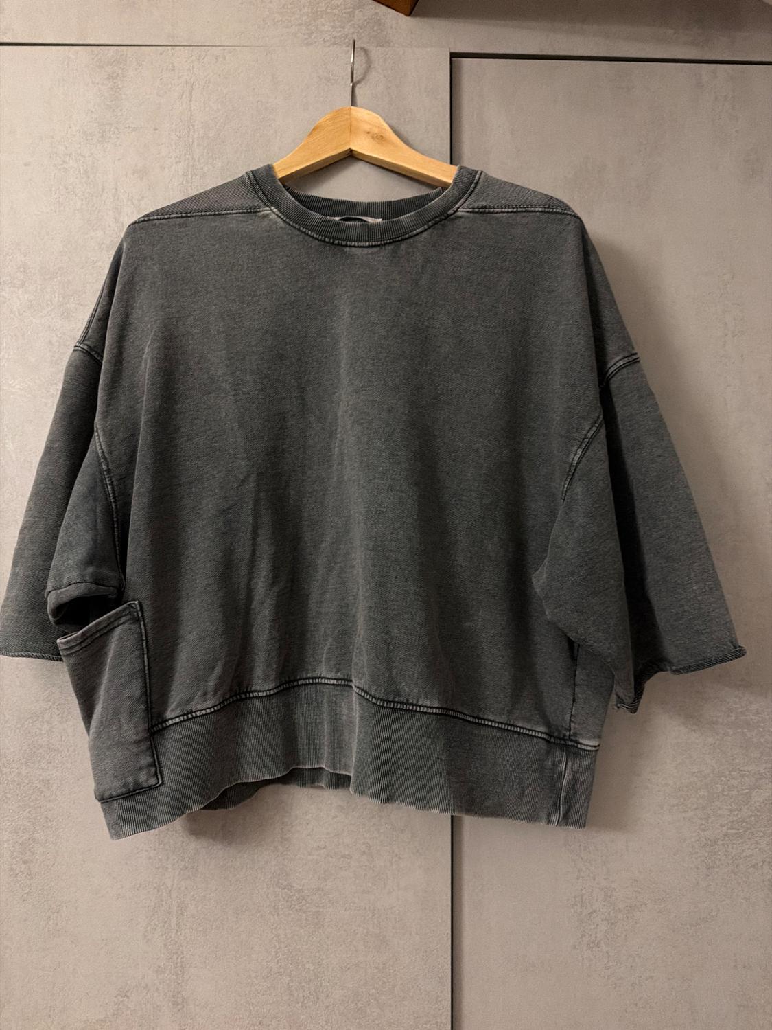 Dark grey casual sweatshirt with a round neck, wide short sleeves, and a side pocket, hanging on a wooden hanger against a grey background.