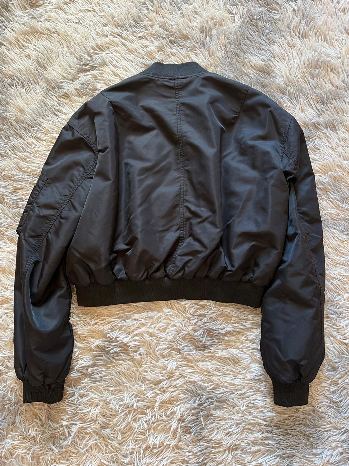Μαύρο bomber jacket με την πλάτη προς τα πάνω, πάνω σε μπεζ χνουδωτό χαλί. Μακριά μανίκια και ελαστικές μανσέτες. Casual και μοντέρνο στυλ.