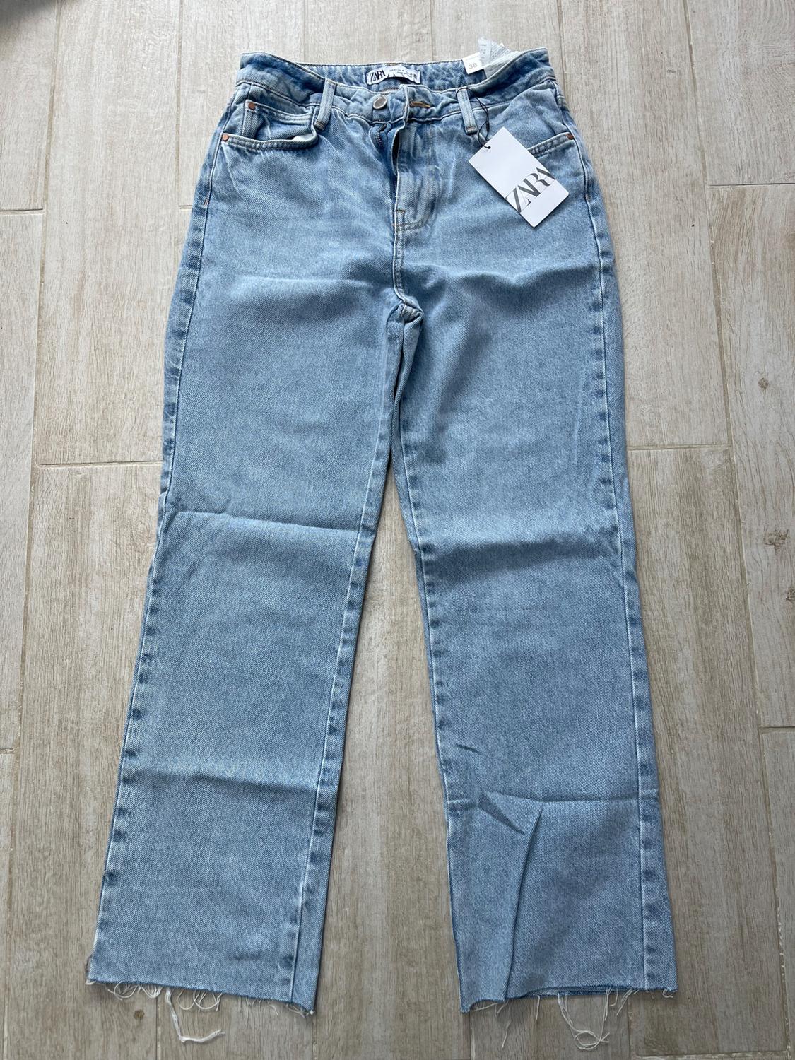 Light blue, straight-leg jeans with a frayed hem, visible Zara label, on light wood flooring.