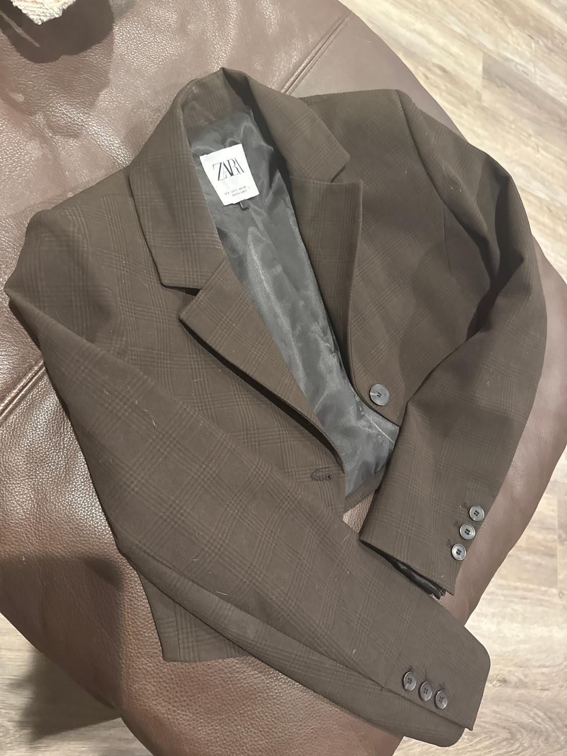 Brown jacket with a plaid pattern, visible Zara tag, on a brown leather sofa. Light wood floor background.