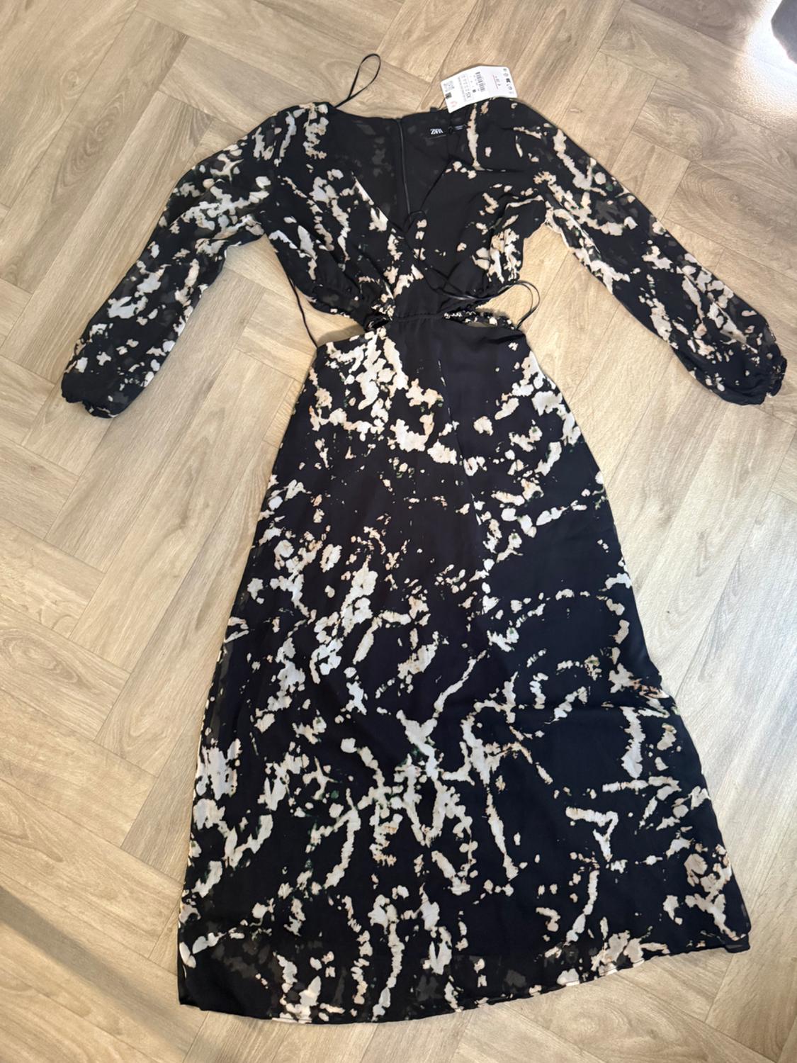 Long black dress with white abstract print, V-neck, long sleeves and cut-out waist, on a wooden floor.