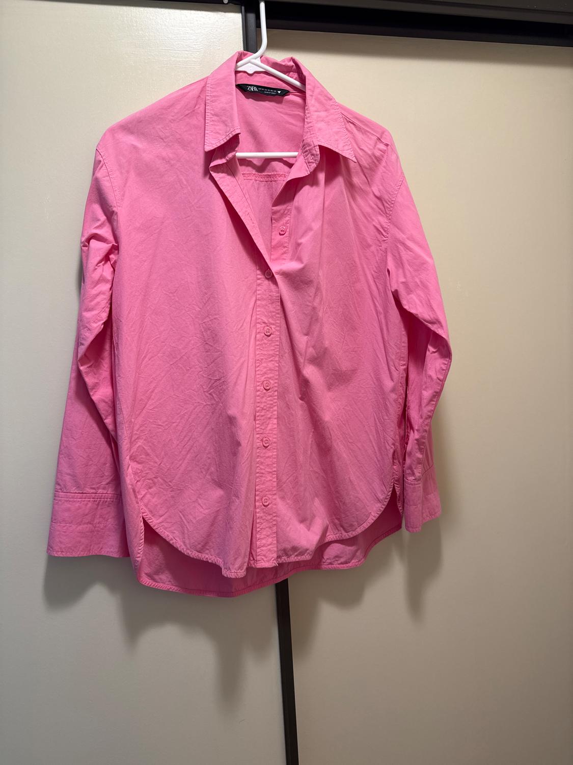 Pink long-sleeved button-down shirt, hanging on a white hanger over a light-colored door. Interior label with the text ZARA.