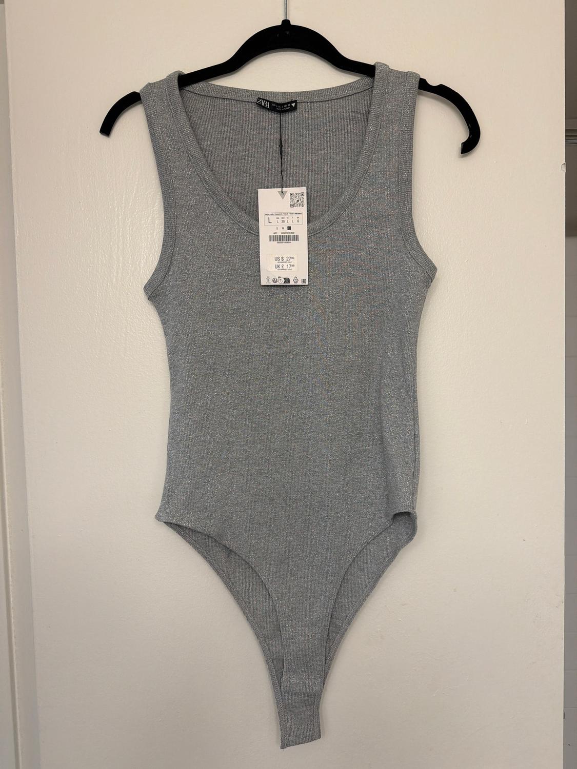Grey, sleeveless bodysuit hanging on a black hanger against a white wall. Visible tag with size and price details.