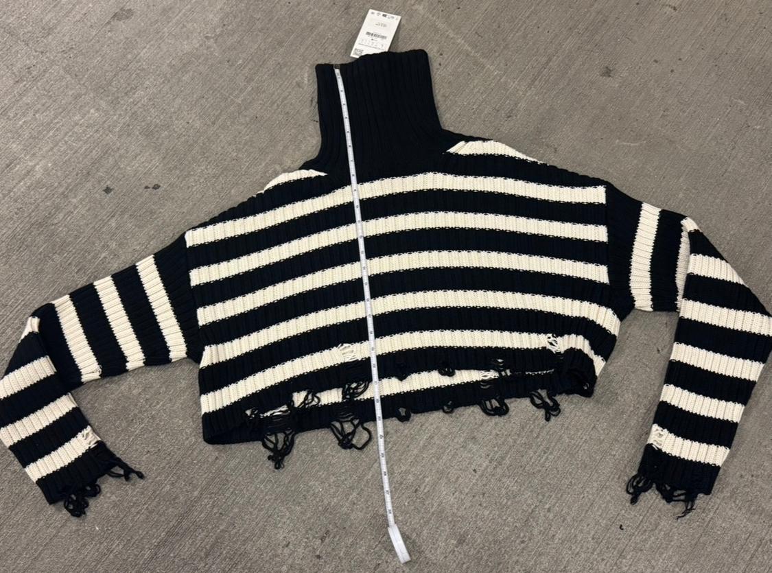Black and white striped turtleneck sweater with frayed edges, laid out on a gray floor with a visible tag.