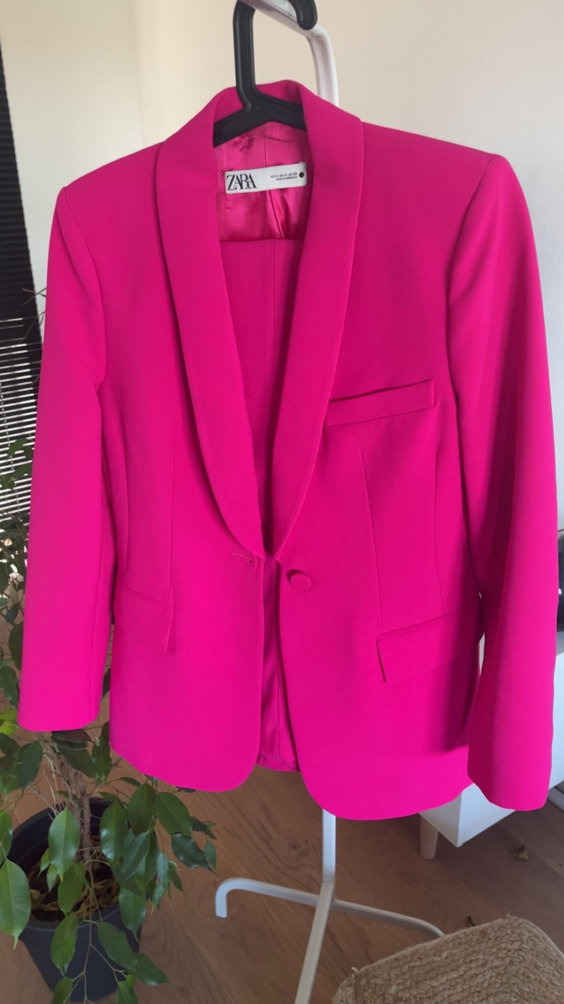 Fuchsia Zara jacket hanging on a black hanger, with a plant and furniture background in a lit room.