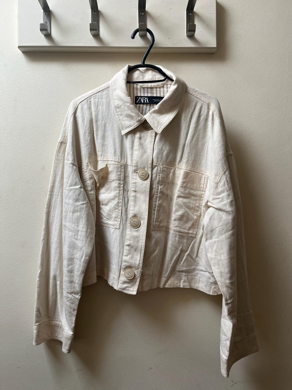 Beige long-sleeved jacket with buttons and pockets, hanging on a wall coat rack. Inner label with the text Zara.